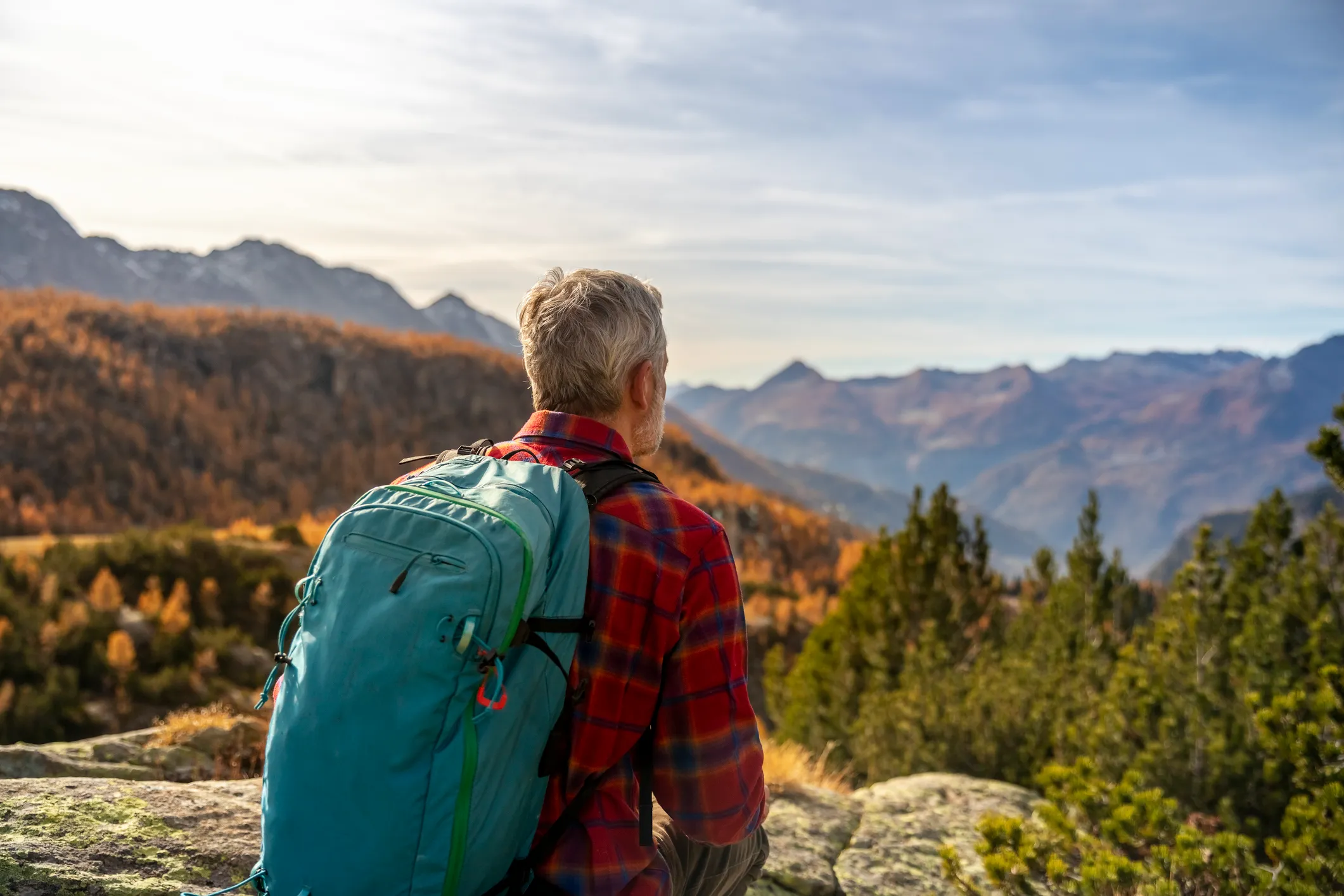 Man with backpack seated in daytime, overlooking mountains.